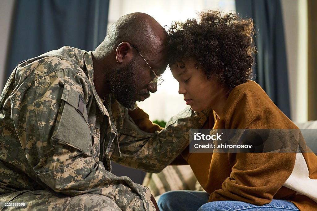 Black middle aged man in military uniform comforting Black young adult woman at home, both sitting close with foreheads touching, showing emotional family support and connection